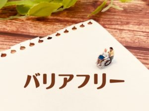 バリアフリーのイメージ。白い紙にバリアフリーのロゴ。車椅子介助をしている人形が置かれている。